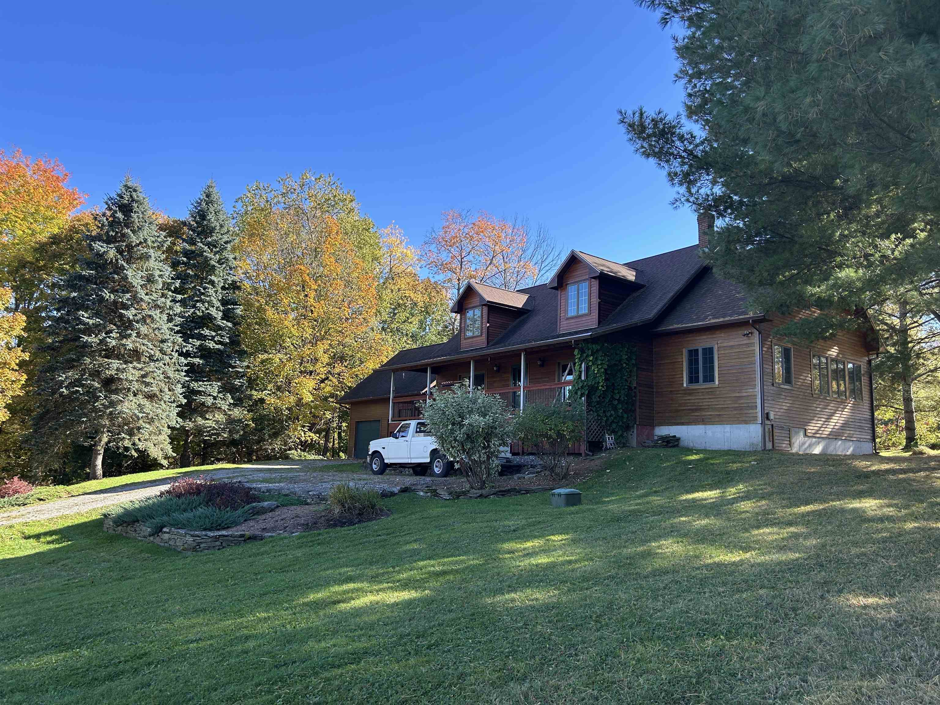 Wood-sided home with dormers and a covered porch, surrounded by autumn trees and a sloping lawn.