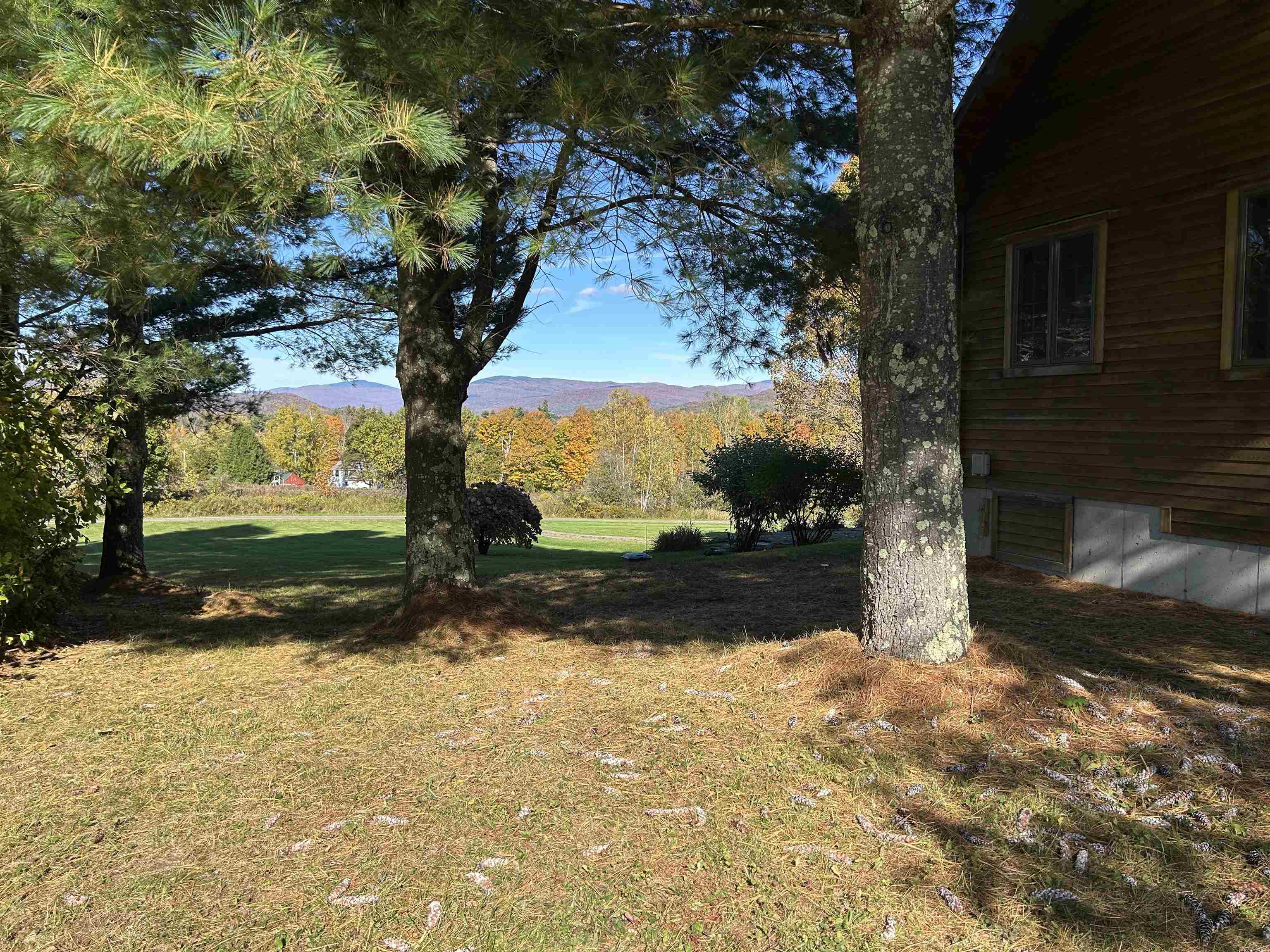 Wood-sided house beside a pine-shaded yard, with autumn foliage and mountains beyond.