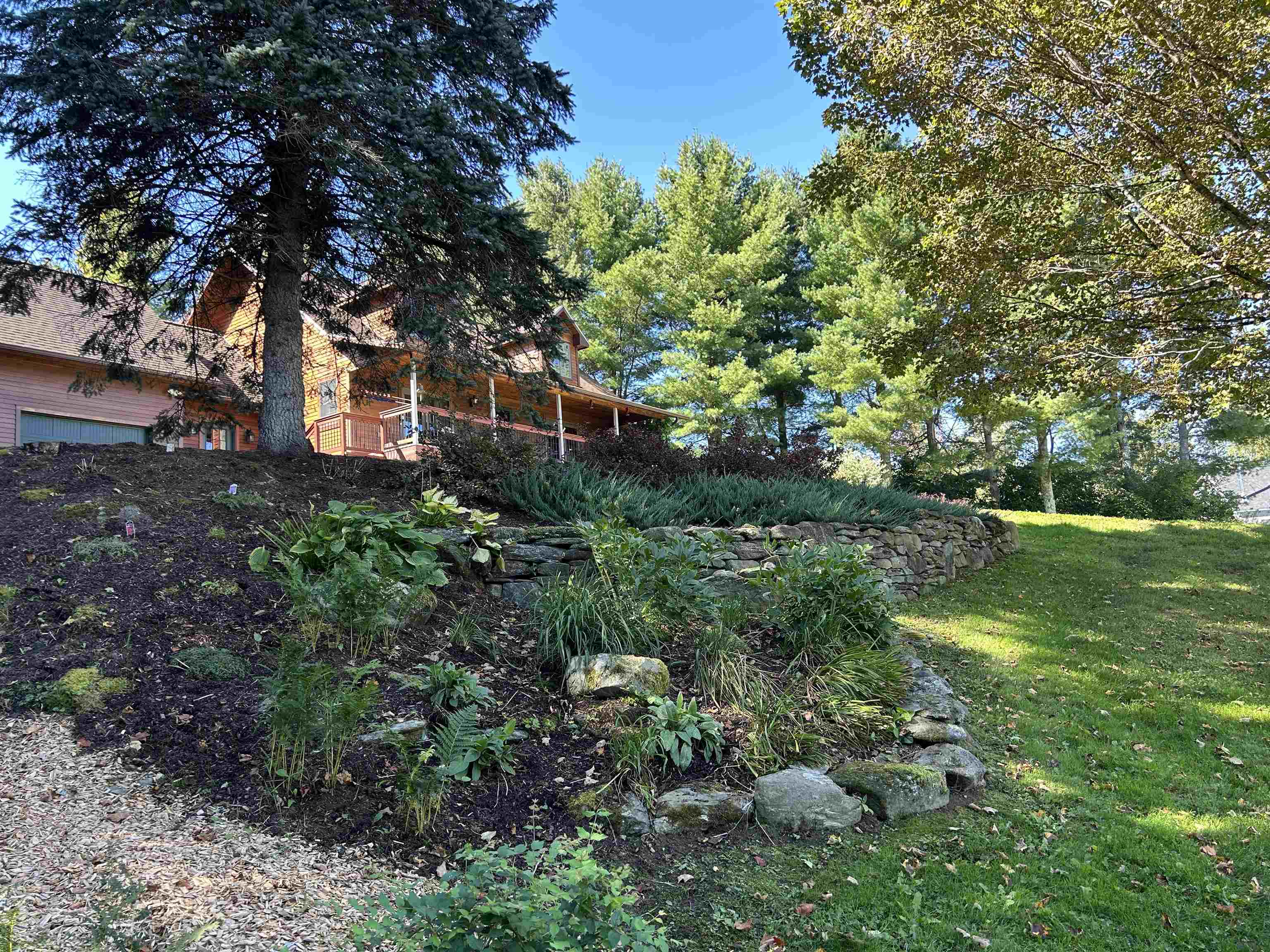 Wood-sided house behind mature trees, above a landscaped garden and stone retaining wall.