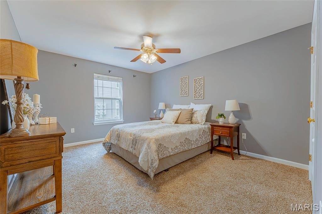 Bedroom with a patterned bed, gray walls, beige carpet, and a wooden ceiling fan.