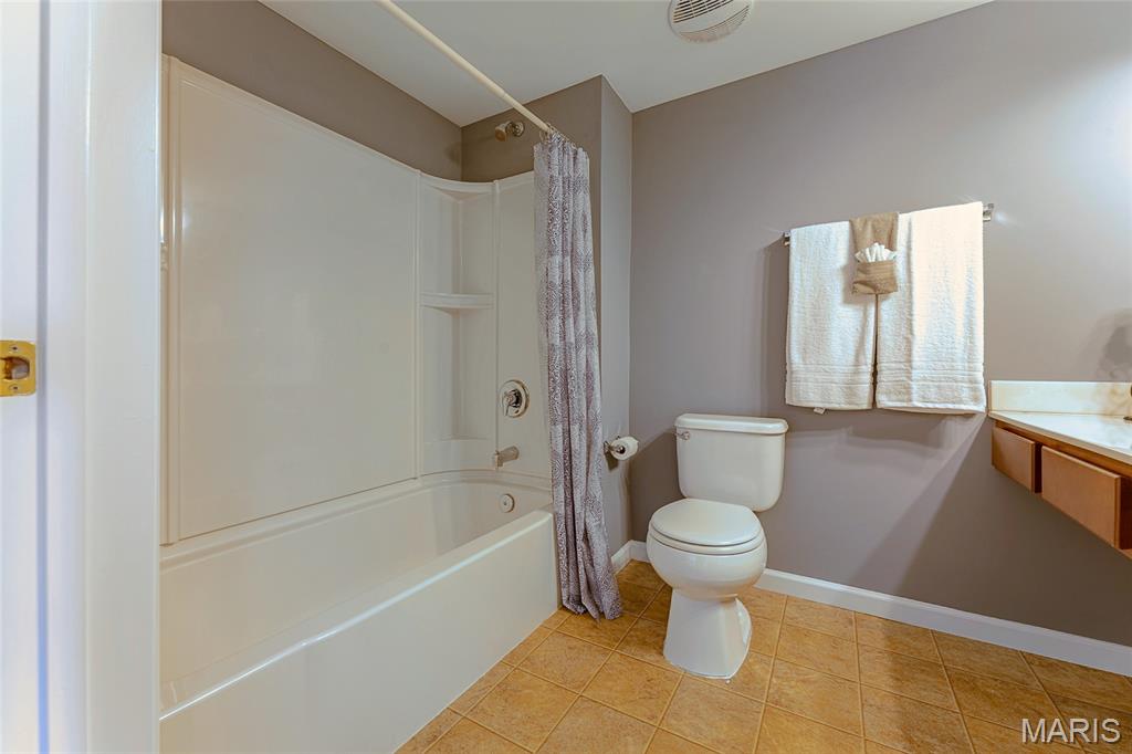 Bathroom with a white tub and shower, toilet, white towels, and tan tile floor.