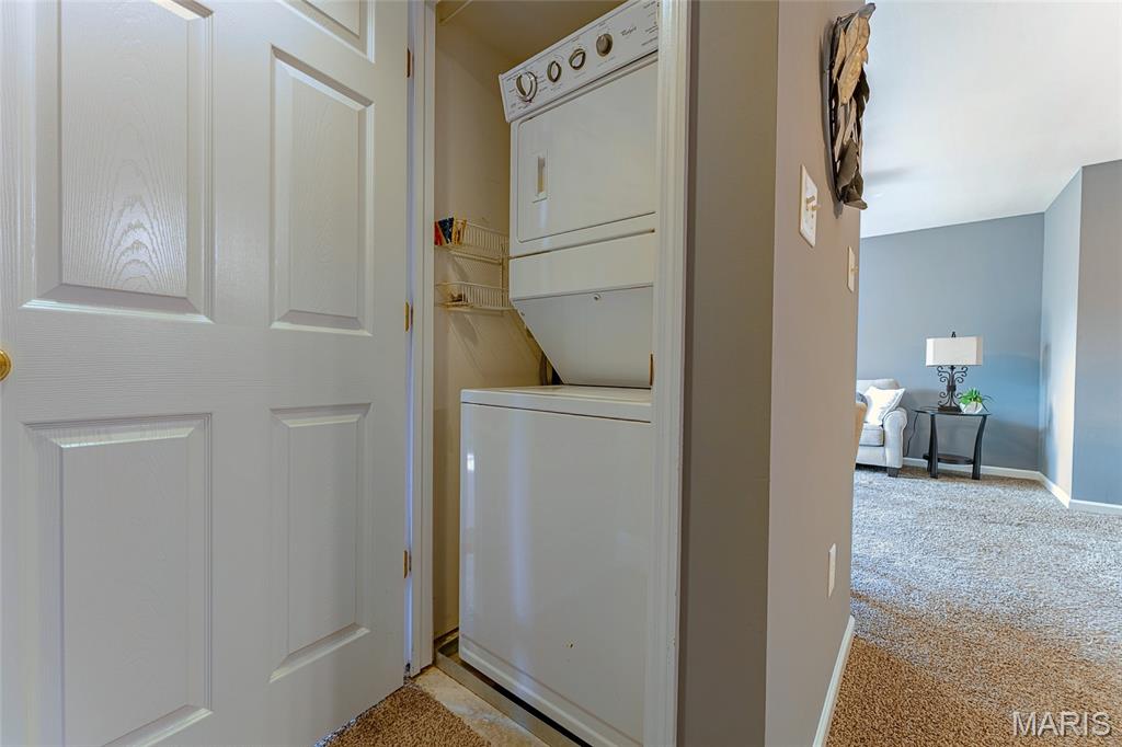 Laundry closet with stacked white washer and dryer beside a carpeted living room.