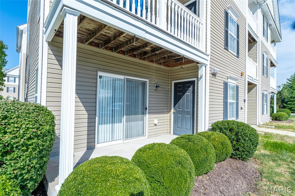 Multi-unit building entrance with a covered balcony, sliding glass door, and neatly trimmed shrubs.
