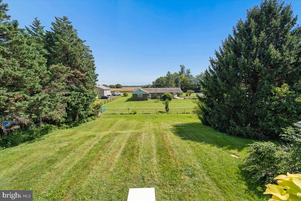 Wide mowed backyard bordered by tall evergreens, with a fenced yard and brick outbuilding beyond.