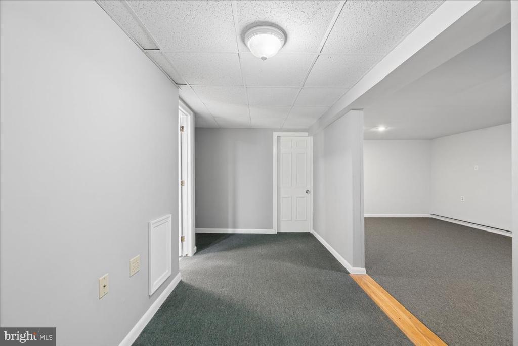Empty carpeted hallway opening into a room, with a white paneled door and wood flooring at the threshold.