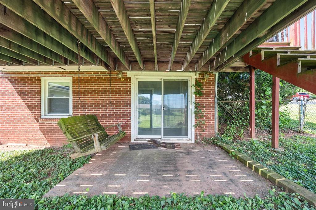 Covered concrete patio beneath a raised deck, with a brick wall, sliding glass door, and weathered bench.