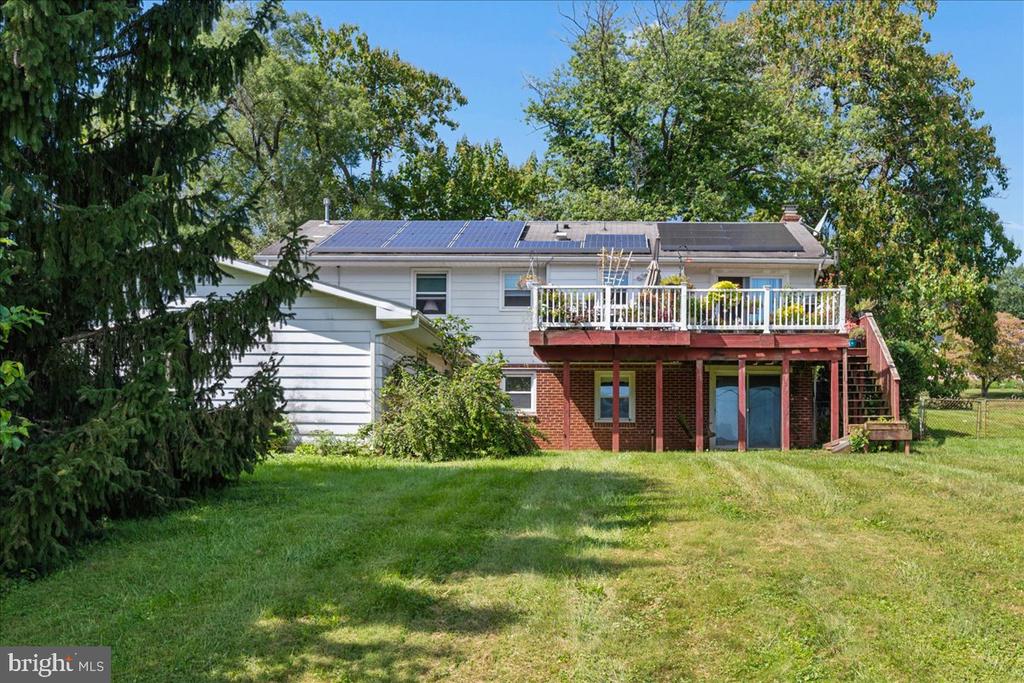 Rear exterior of a two-story house with rooftop solar panels and a raised deck overlooking a broad lawn.