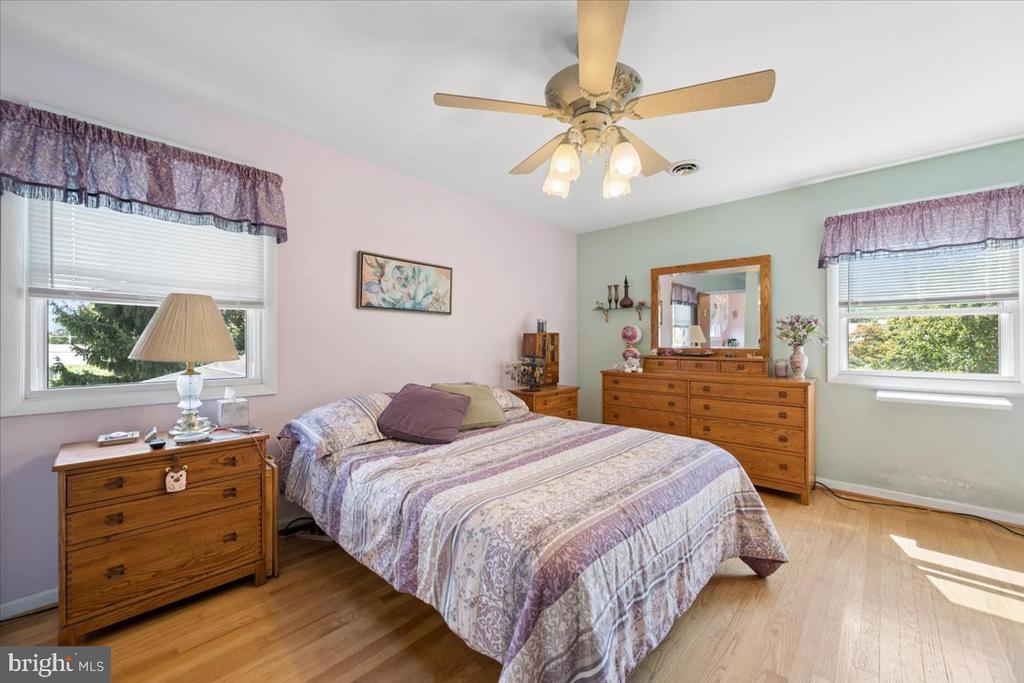 Bedroom with a patterned quilt, wooden dressers, two windows, and a ceiling fan.