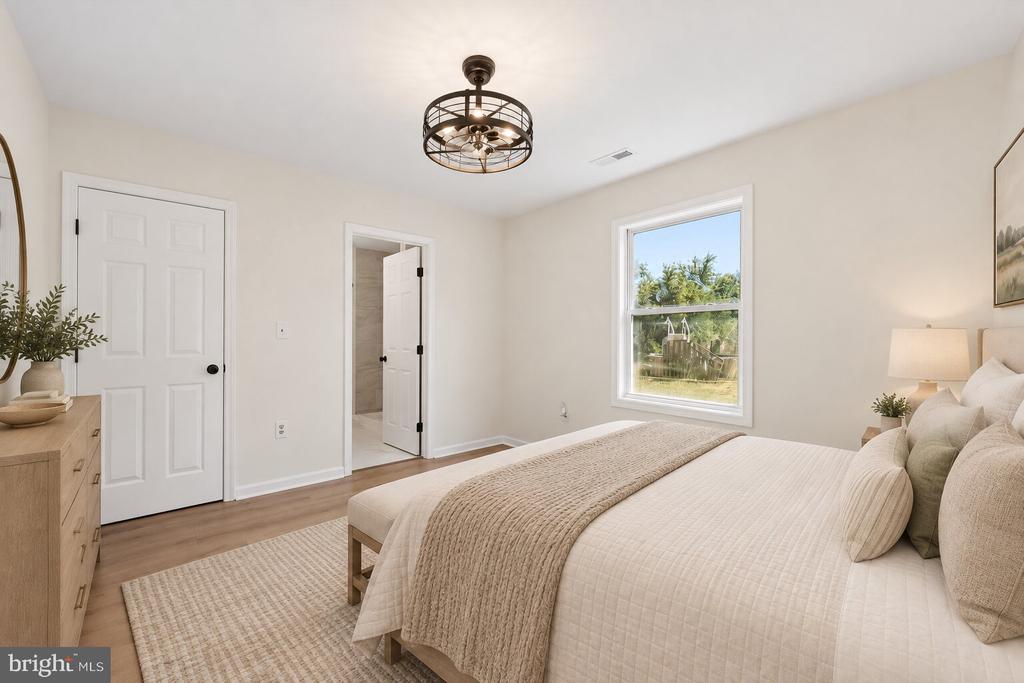 Staged bedroom with neutral bedding, a wood dresser, and a window overlooking greenery.