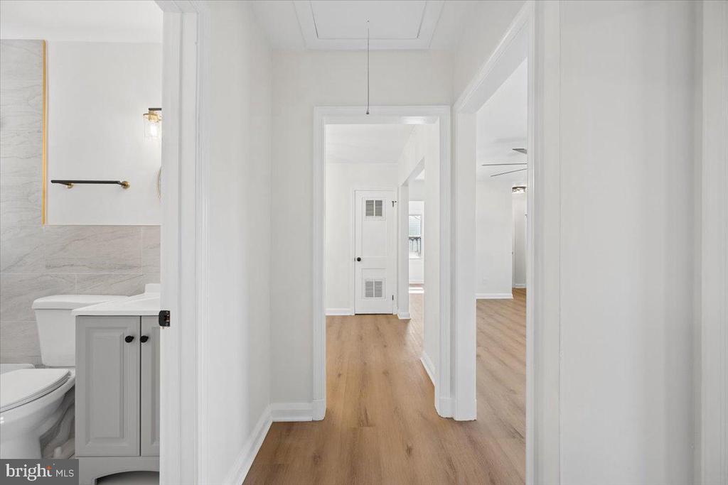 Bright hallway with light wood-look floors, showing a bathroom on the left and adjoining rooms beyond.