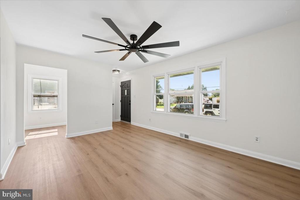 Vacant living room with light wood floors, large street-facing windows, and a dark ceiling fan.