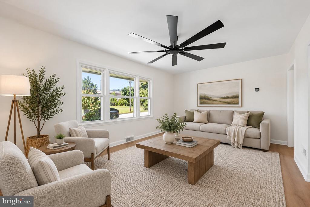 Staged living room with beige seating, large windows, and a black ceiling fan.