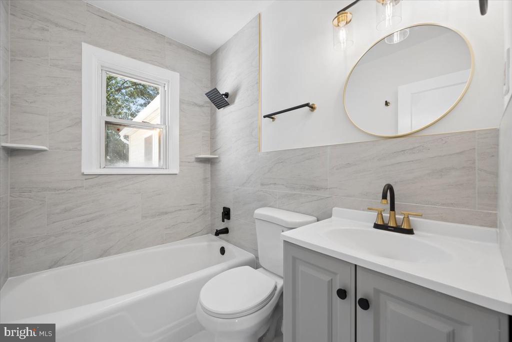 Bathroom with gray tile, white tub, and gray vanity beneath a round gold-framed mirror.