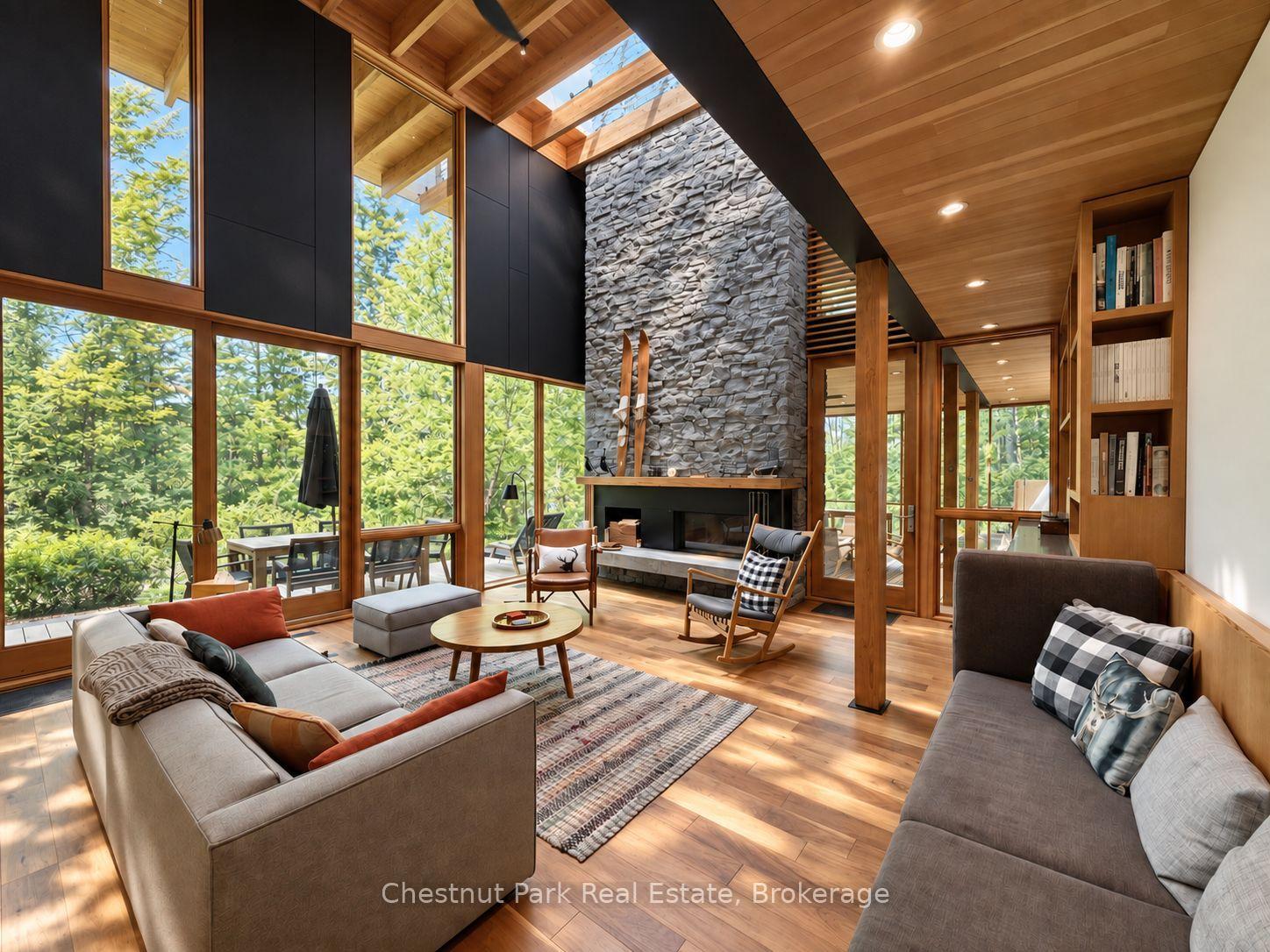 Living room with a tall stone fireplace, timber ceiling, skylights, and expansive windows overlooking trees.