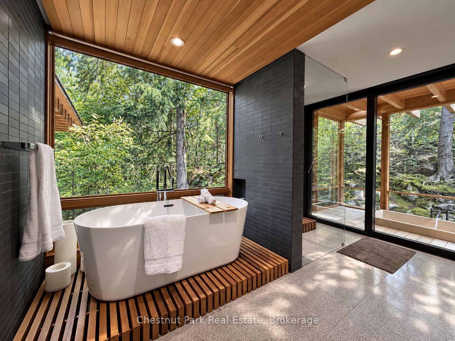 Modern bathroom with a freestanding tub, wood-slat platform, and large windows overlooking trees.