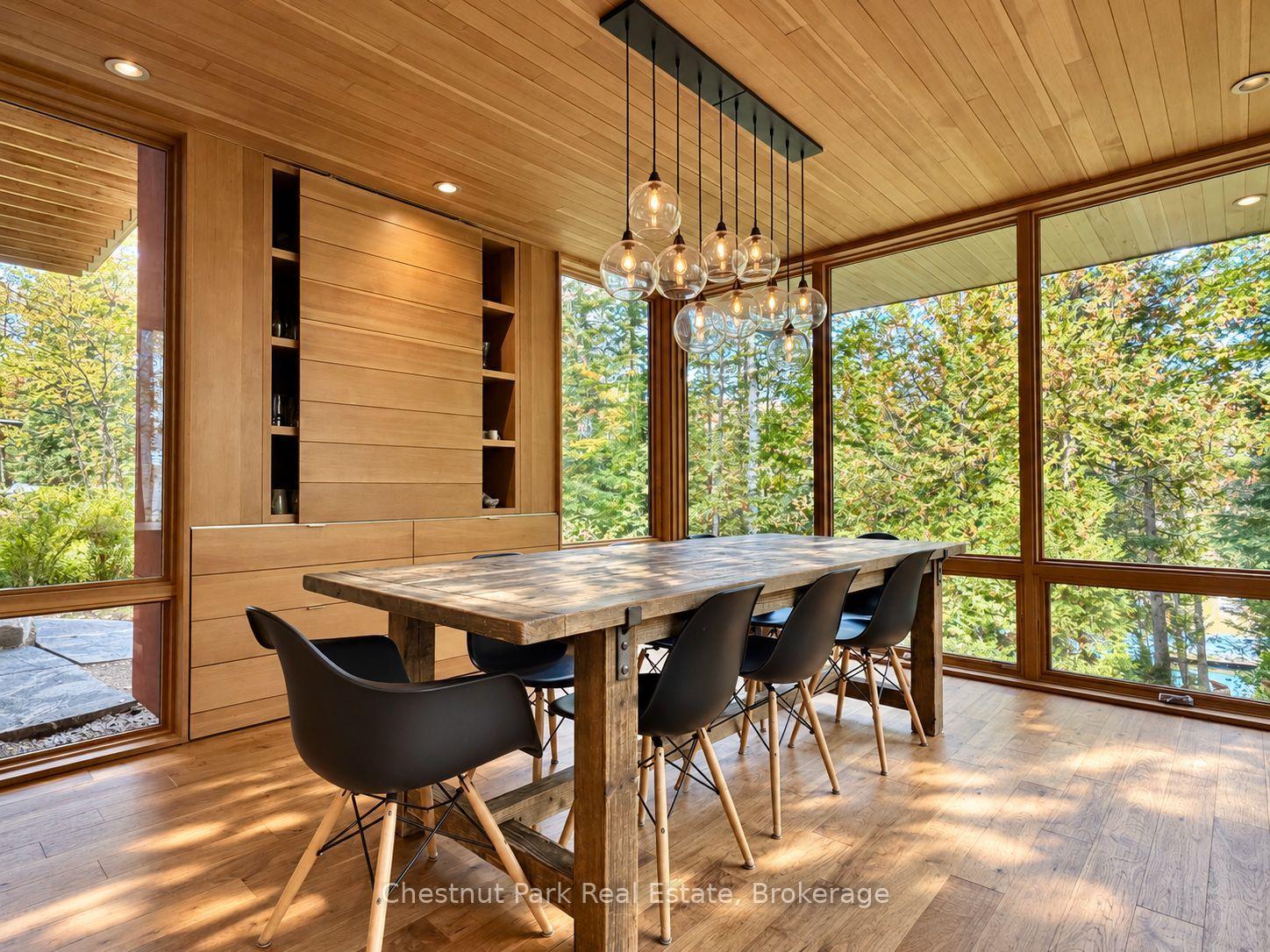 Dining room with a rustic wood table, glass pendant lights, and floor-to-ceiling windows overlooking trees.