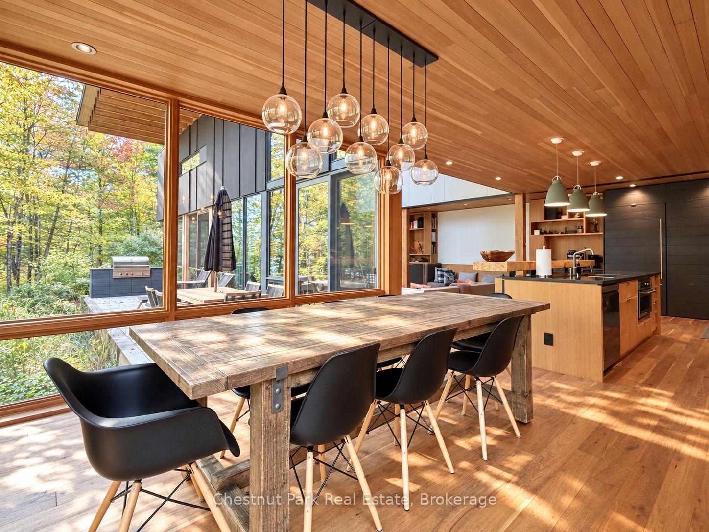 Dining area with a rustic wood table, vaulted wood ceiling, and floor-to-ceiling windows overlooking trees.