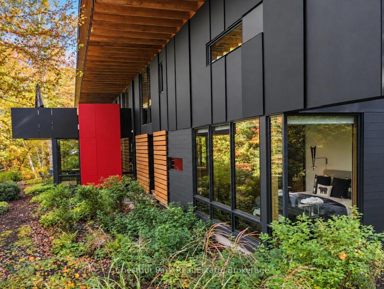 Modern home exterior with dark cladding, a red accent wall, and a wood-covered overhang amid autumn foliage.
