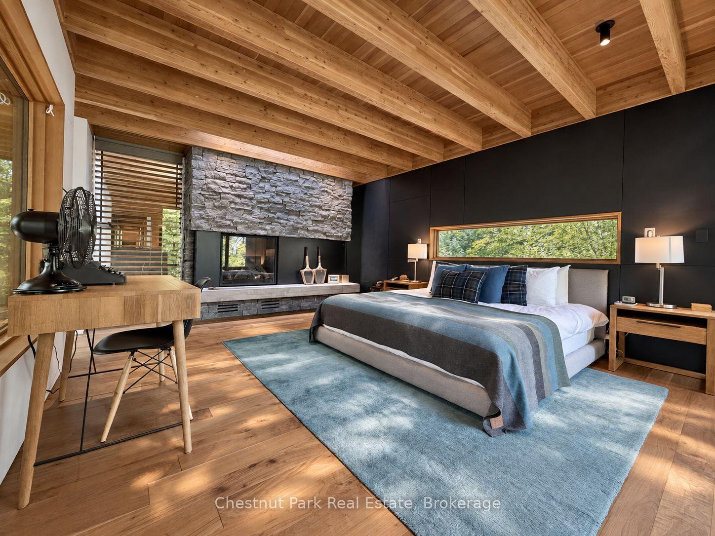 Modern bedroom with exposed timber ceiling, stone fireplace, and a large bed on a blue rug.