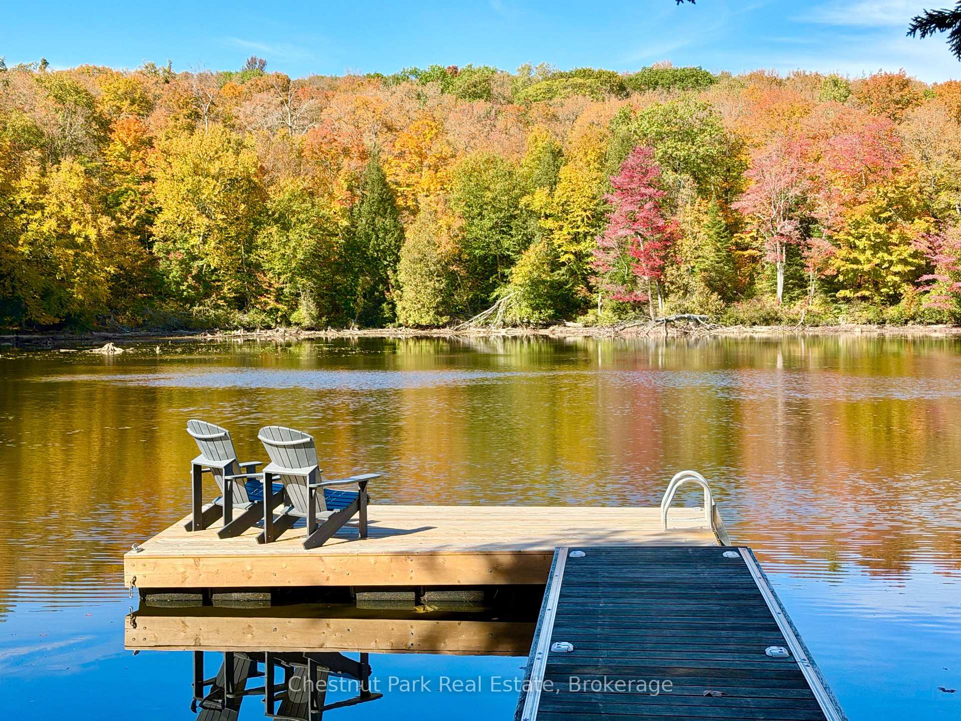 Wooden lake dock with two Adirondack chairs, backed by colorful autumn trees.