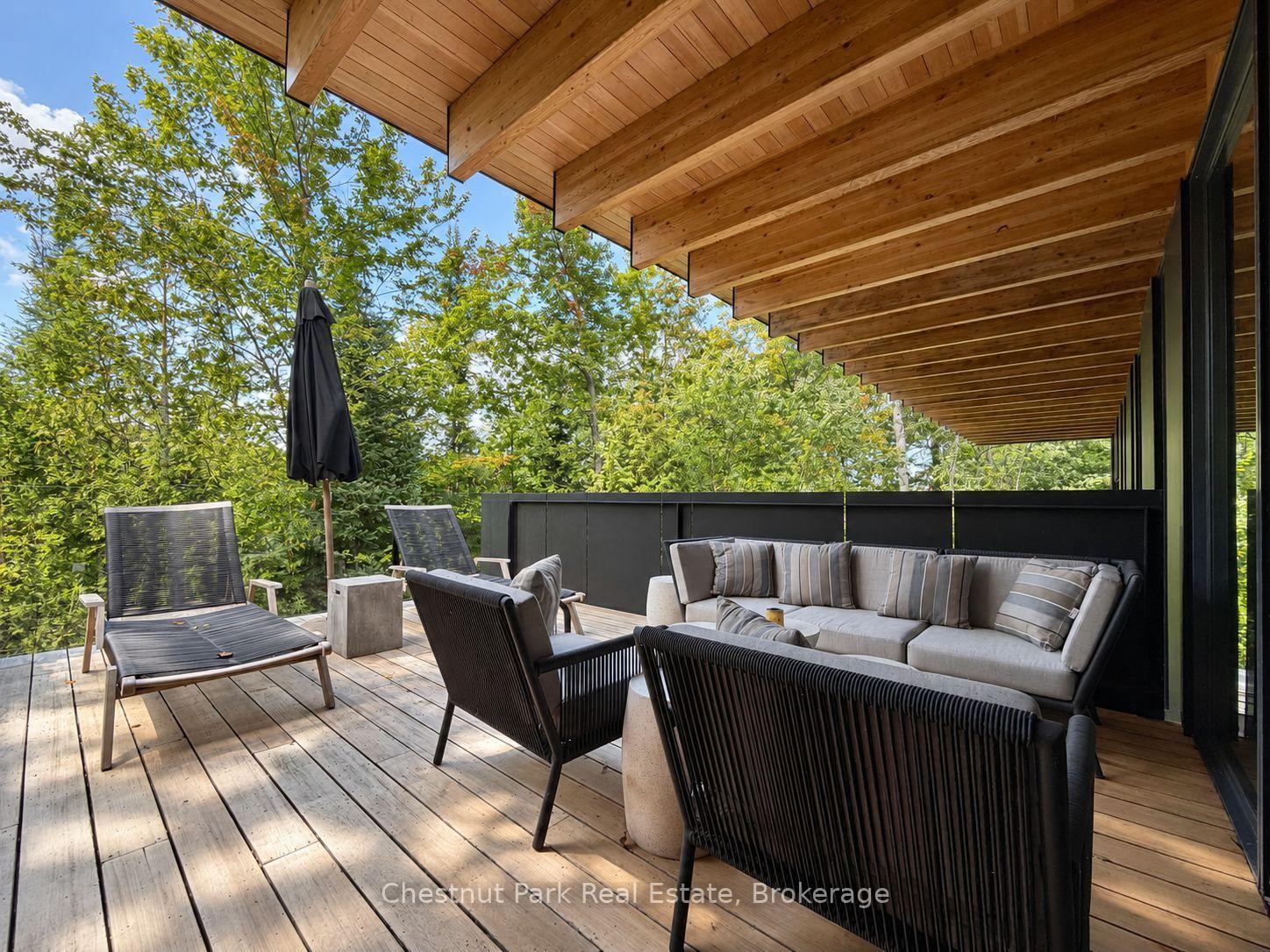 Wooden deck with a covered lounge area, woven chairs, and a cushioned sofa overlooking leafy trees.