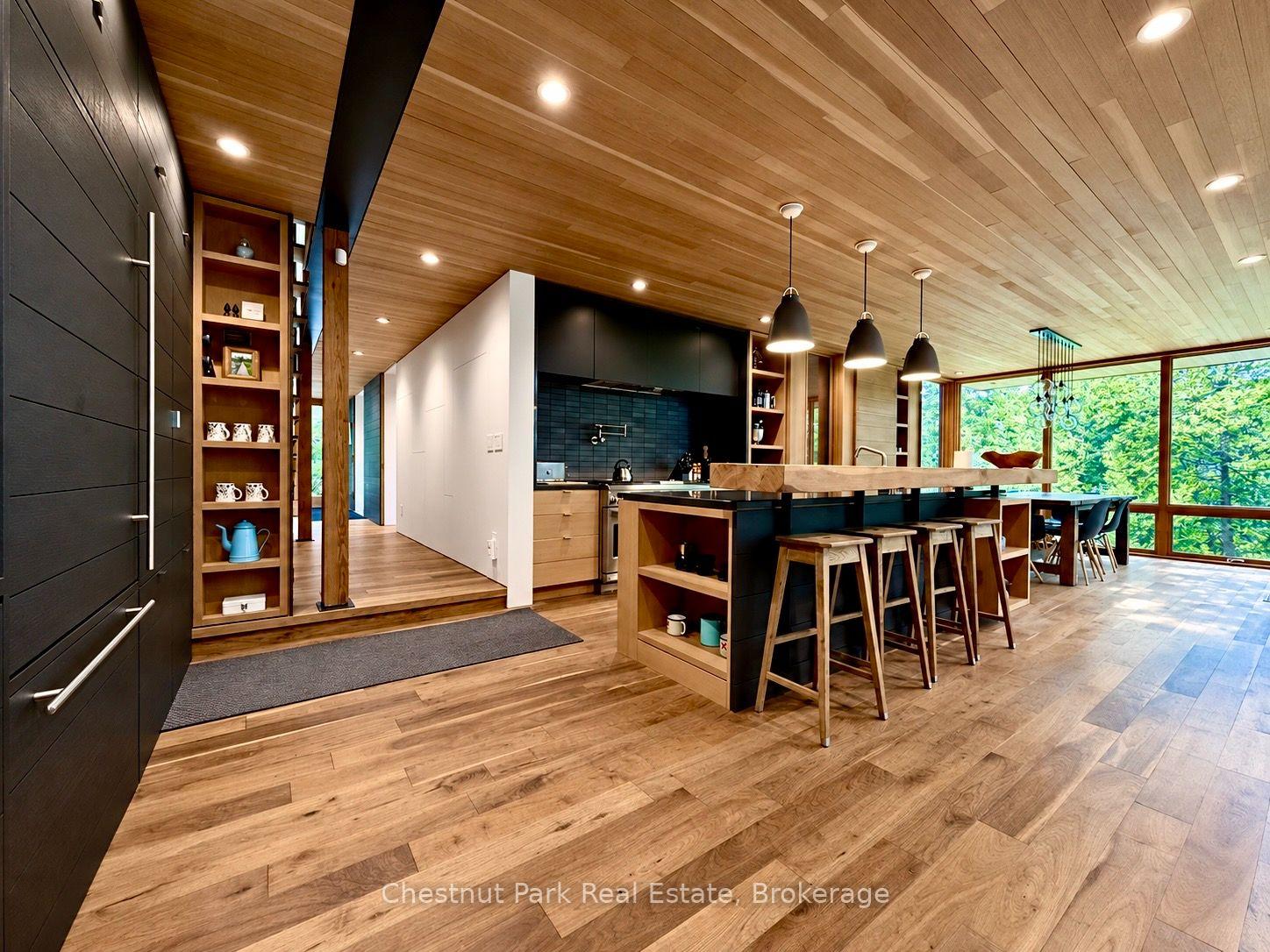 Modern kitchen with a black island, wood countertop, bar stools, and floor-to-ceiling windows overlooking trees.