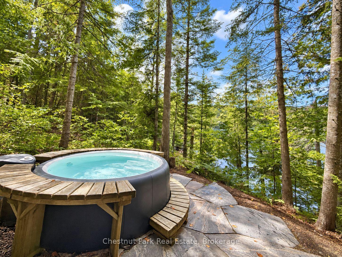 Outdoor hot tub on a circular wooden deck, surrounded by trees with a glimpse of water beyond.
