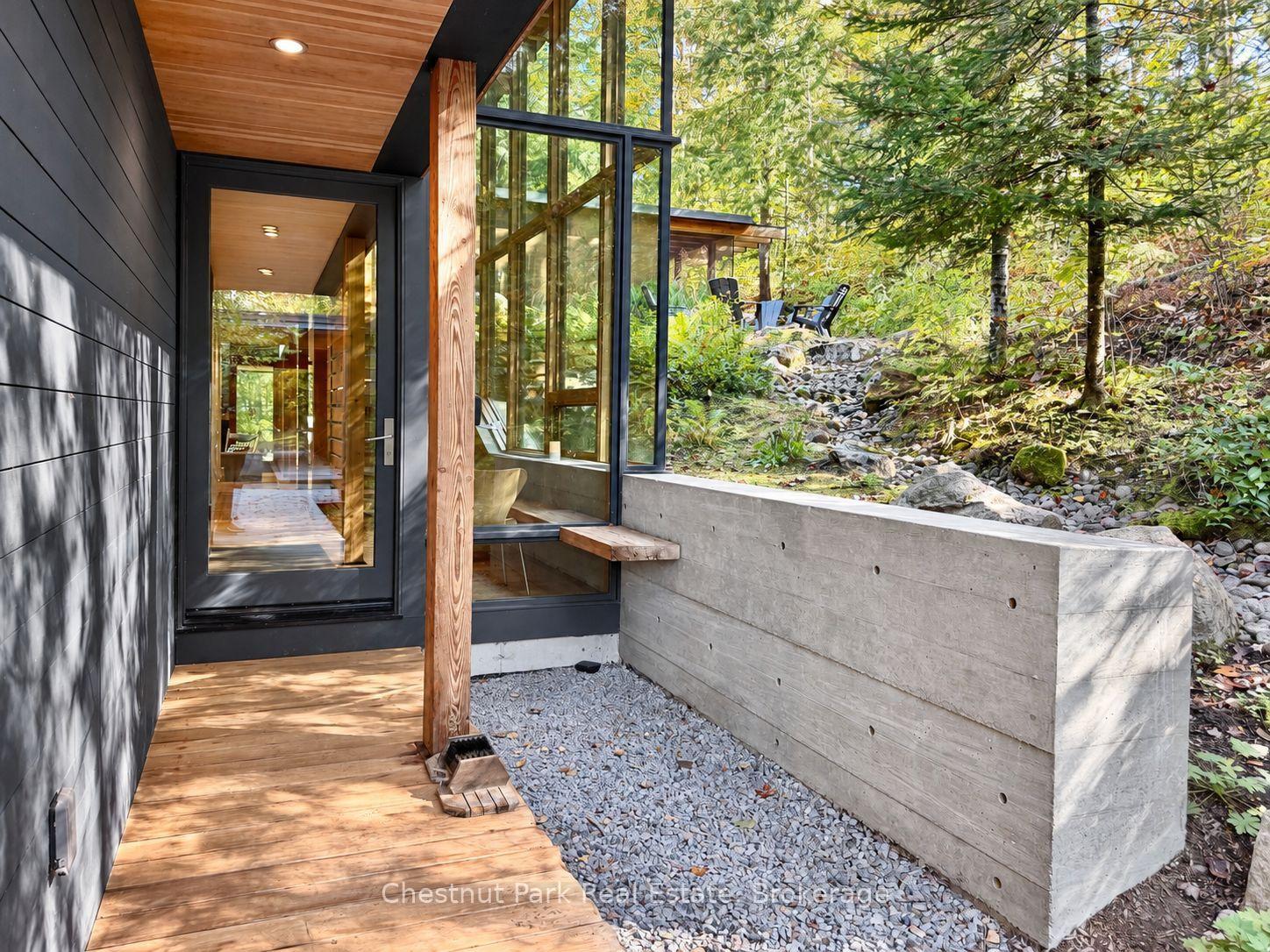 Modern home entrance with a wood-plank porch, tall glass windows, and a wooded rocky slope.