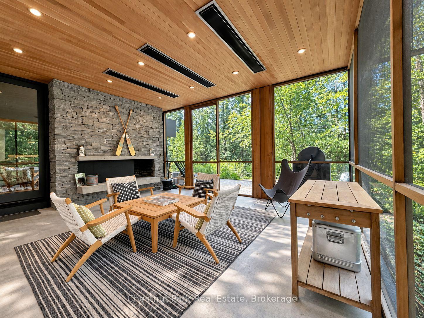 Sunroom with a stone fireplace, wood ceiling, and expansive windows overlooking trees.