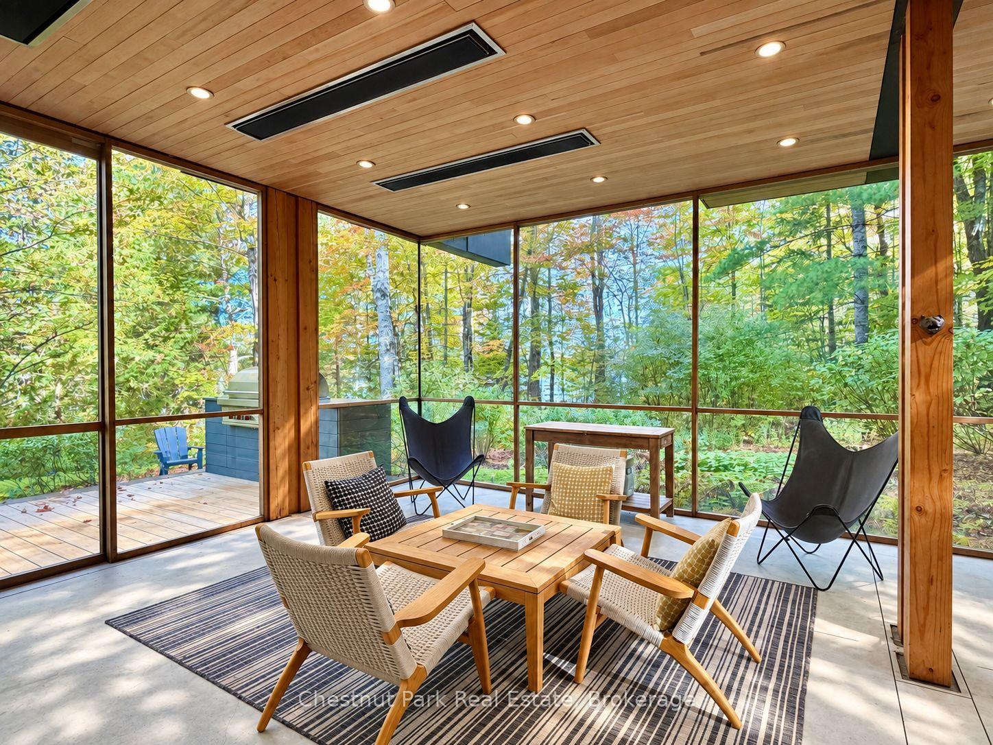 Glass-walled sunroom with wicker seating and a wood ceiling overlooking leafy woods.