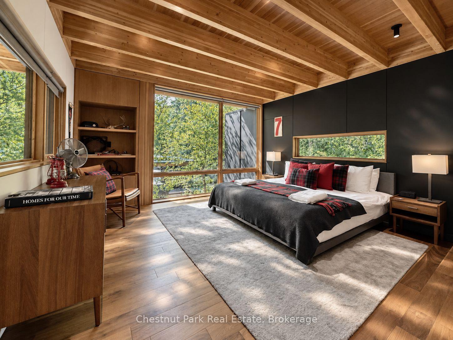 Bedroom with an exposed timber ceiling, dark accent wall, and large windows overlooking trees.