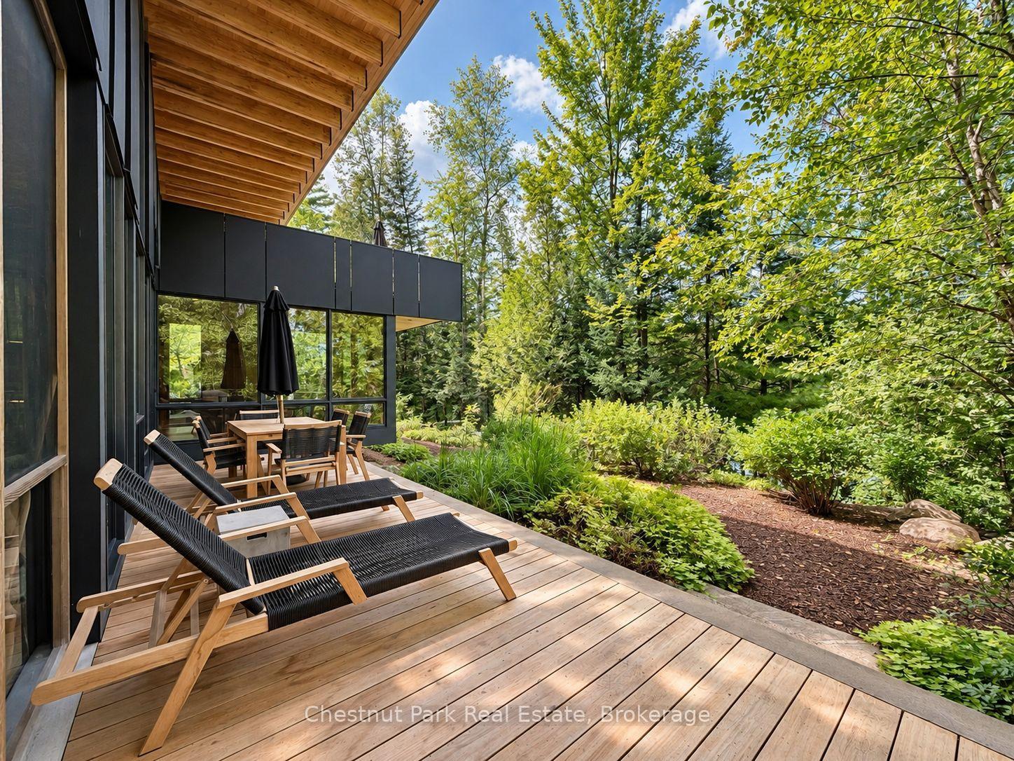 Wooden deck with lounge chairs and dining set beside a modern home, overlooking a wooded garden.