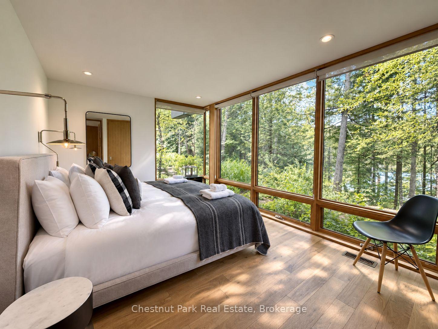 Staged bedroom with floor-to-ceiling windows overlooking leafy trees and glimpses of water.