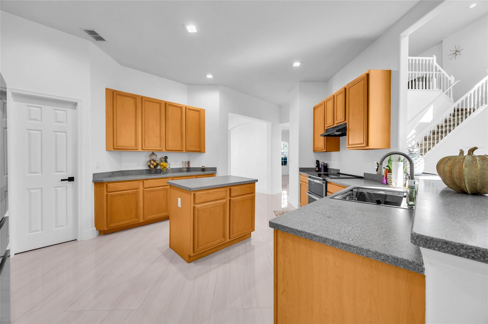 Kitchen with honey-toned wood cabinets, gray counters, a central island, and a staircase beyond.