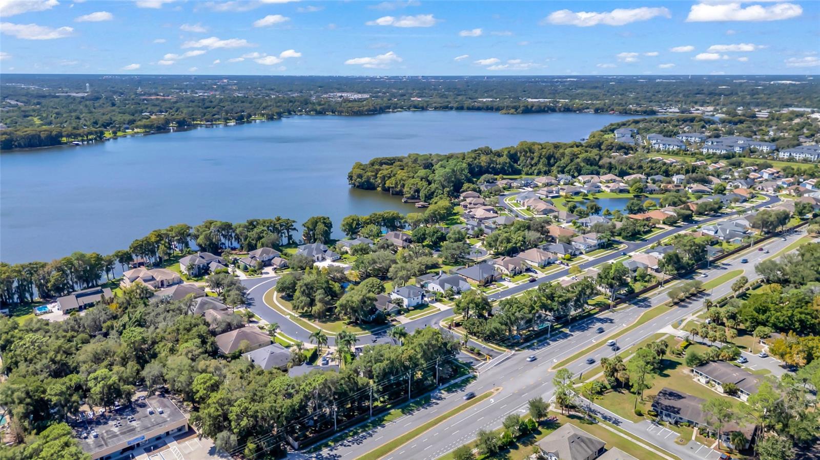 Aerial view of a lakeside neighborhood, with homes, tree-lined shore, and a broad road.