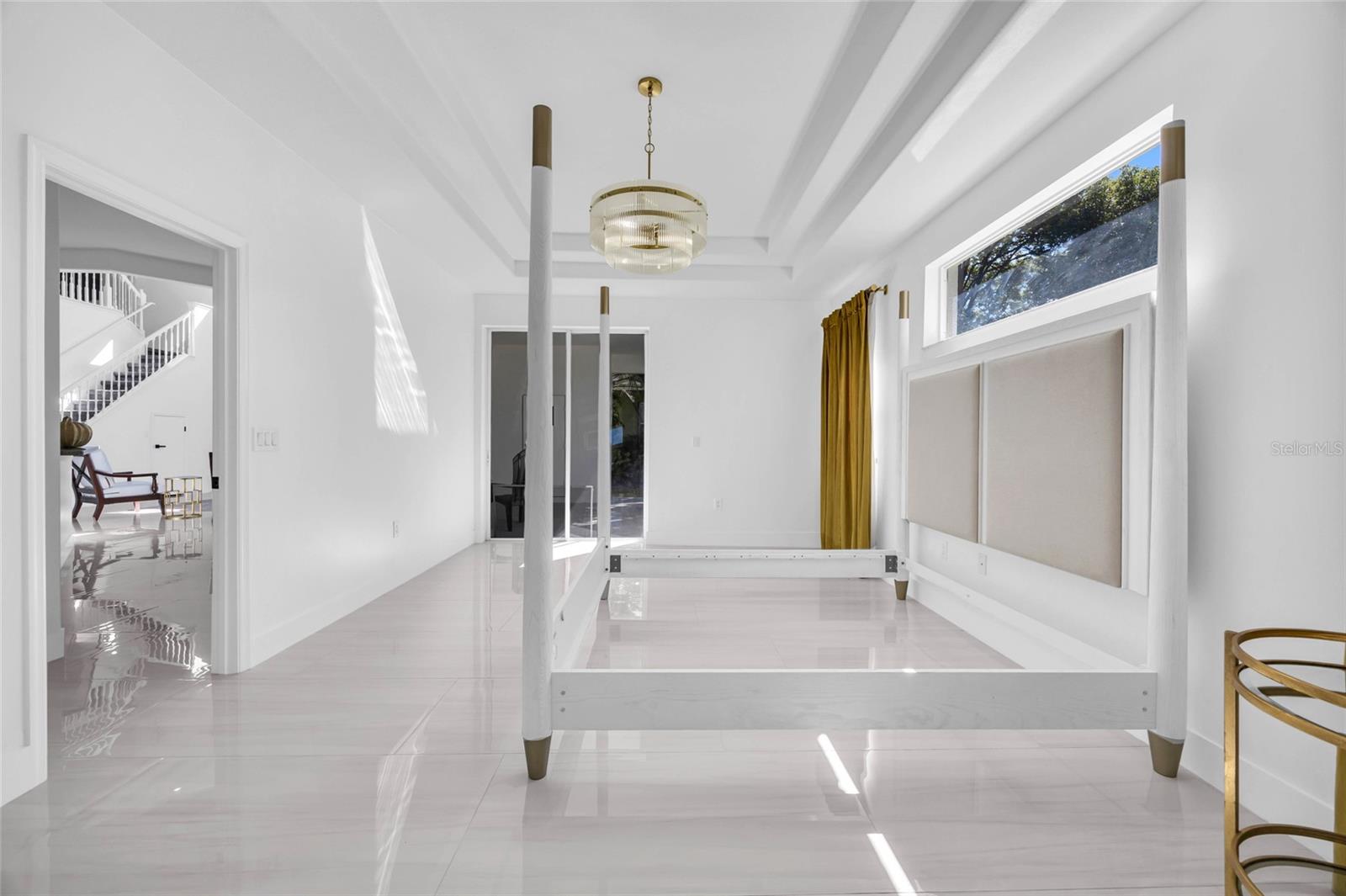 Bright bedroom with a white four-poster bed frame, glossy tile floors, and large windows.