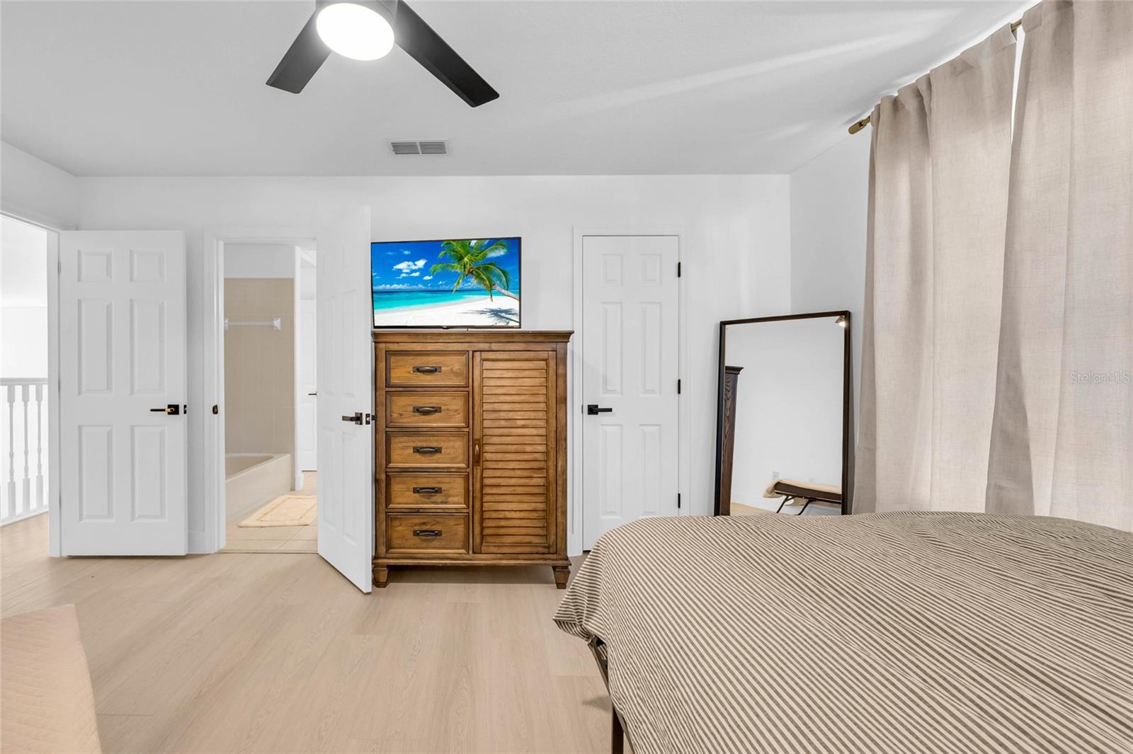 Furnished bedroom with striped bedspread, wooden dresser, ceiling fan, and open bathroom doorway.