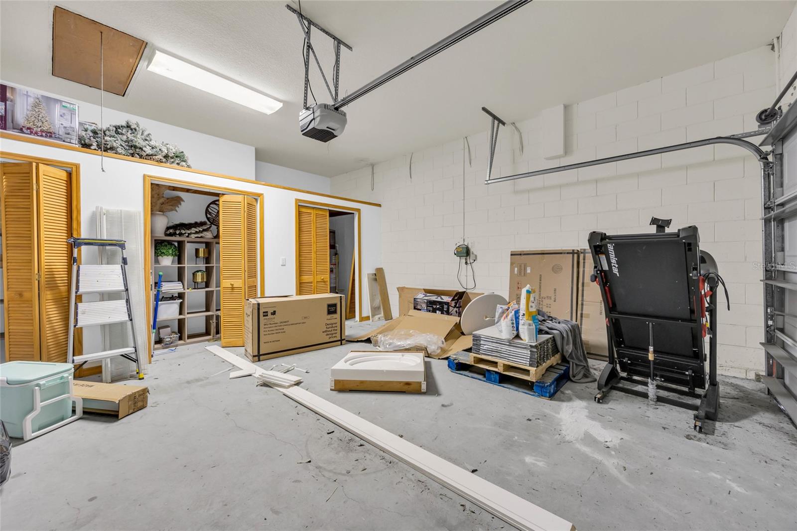 Garage with yellow louvered storage doors, boxes, building materials, and a treadmill.