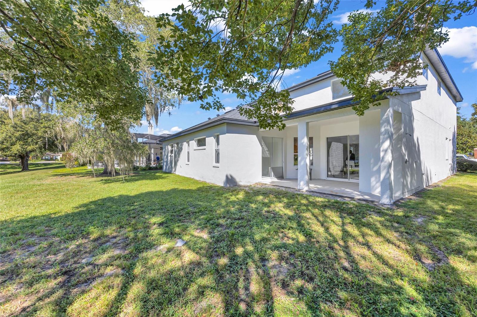 White two-story home with a covered patio and sliding glass doors beneath mature trees.
