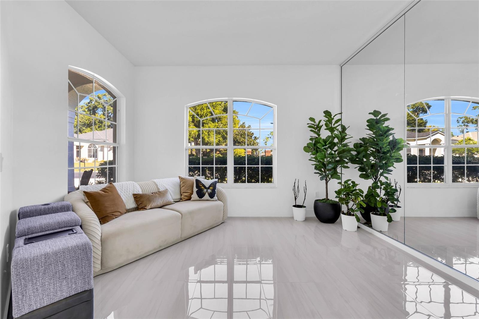 Bright living room with a cream sectional, arched windows, glossy tile floors, and potted plants beside a mirrored wall.