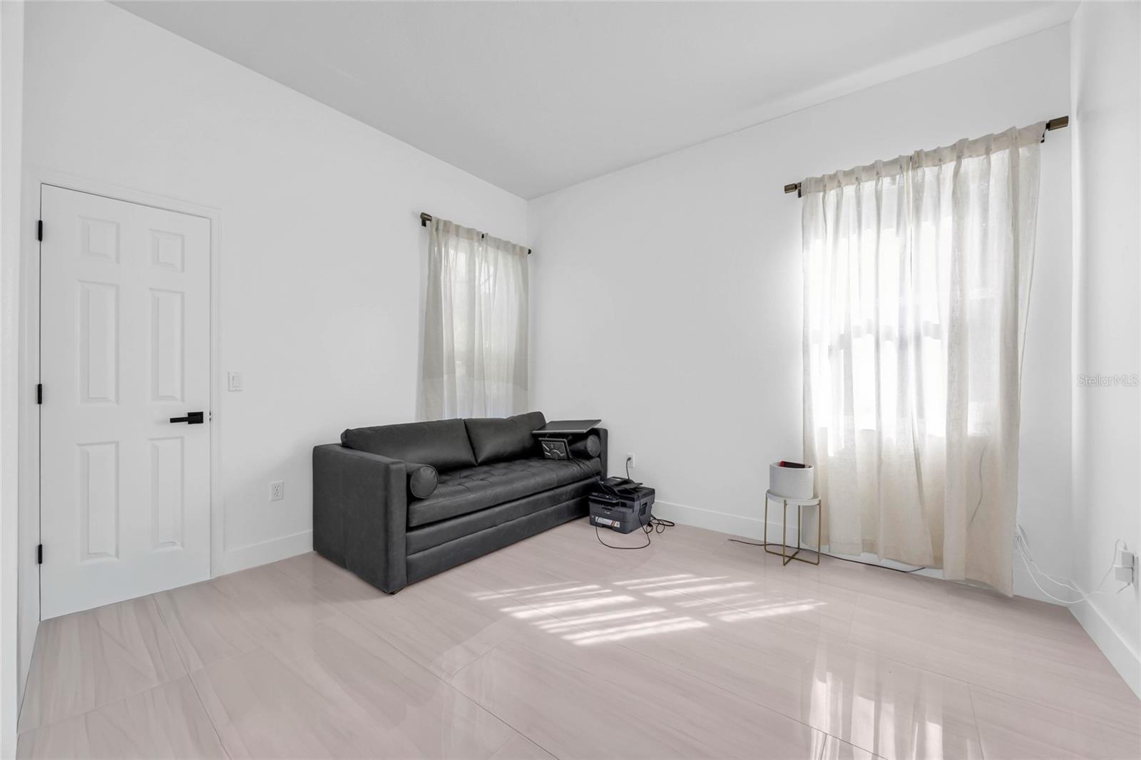 Bright living room with a dark gray sofa, pale tile floors, and sheer-curtained windows.