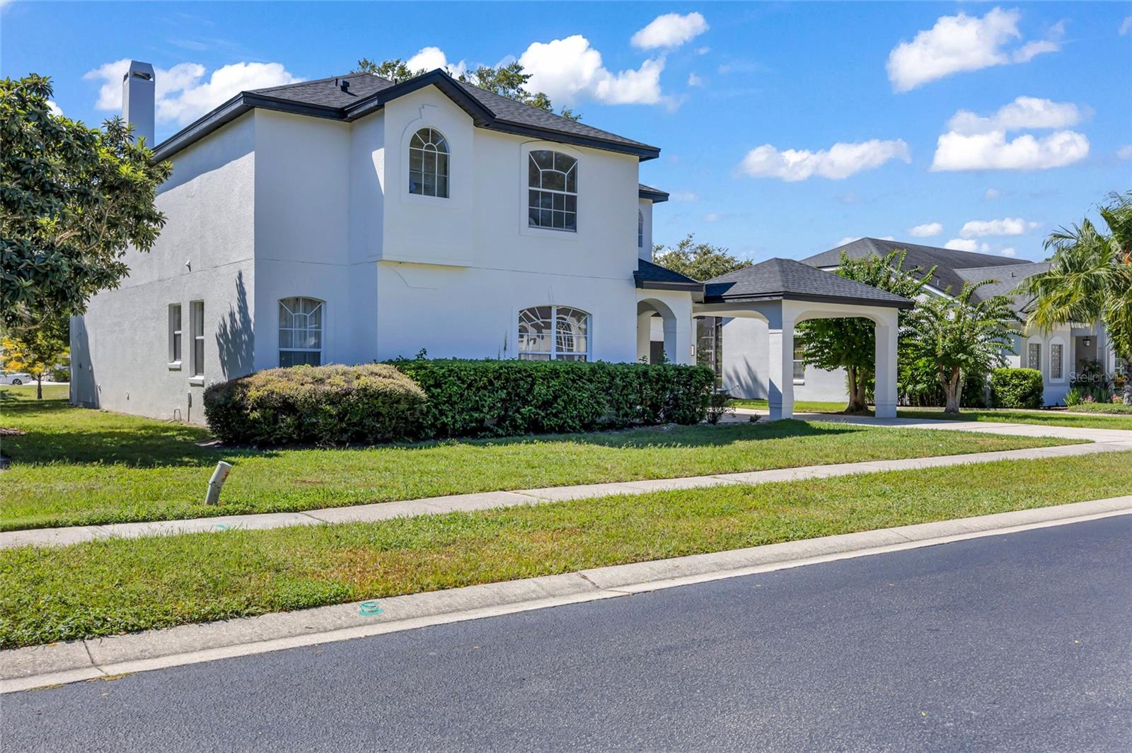Two-story white stucco house with arched windows, trimmed hedges, and a covered driveway entrance.