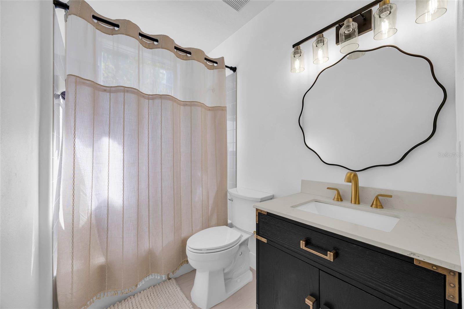 Bathroom with a dark vanity, gold-tone faucet, wavy mirror, and beige shower curtain.