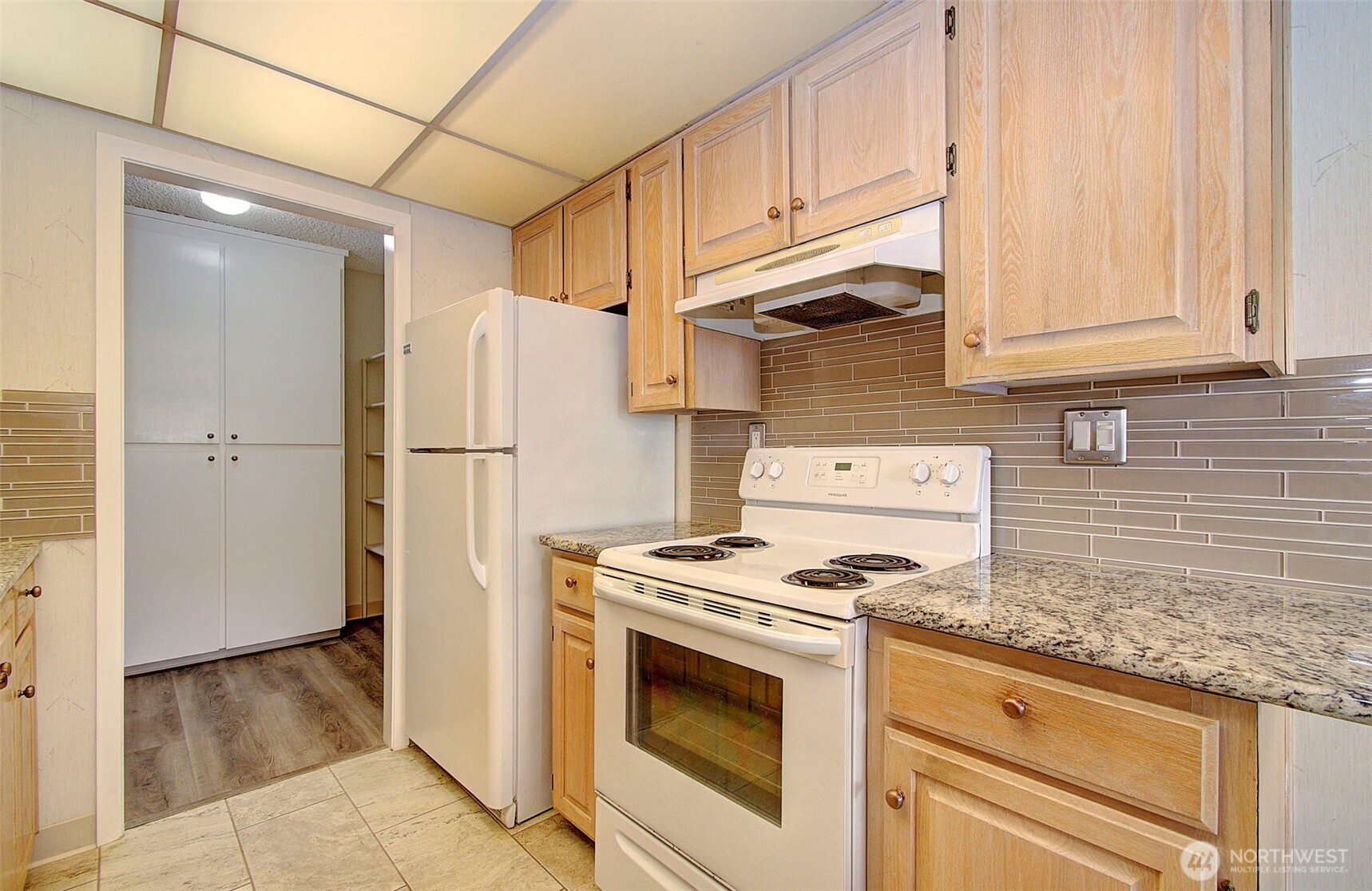 Kitchen with light wood cabinets, granite countertops, and white appliances; pantry cabinets are visible through the doorway.