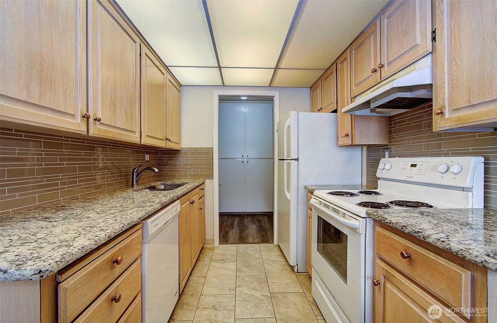 Galley kitchen with light wood cabinets, granite counters, tiled backsplash, and white appliances.