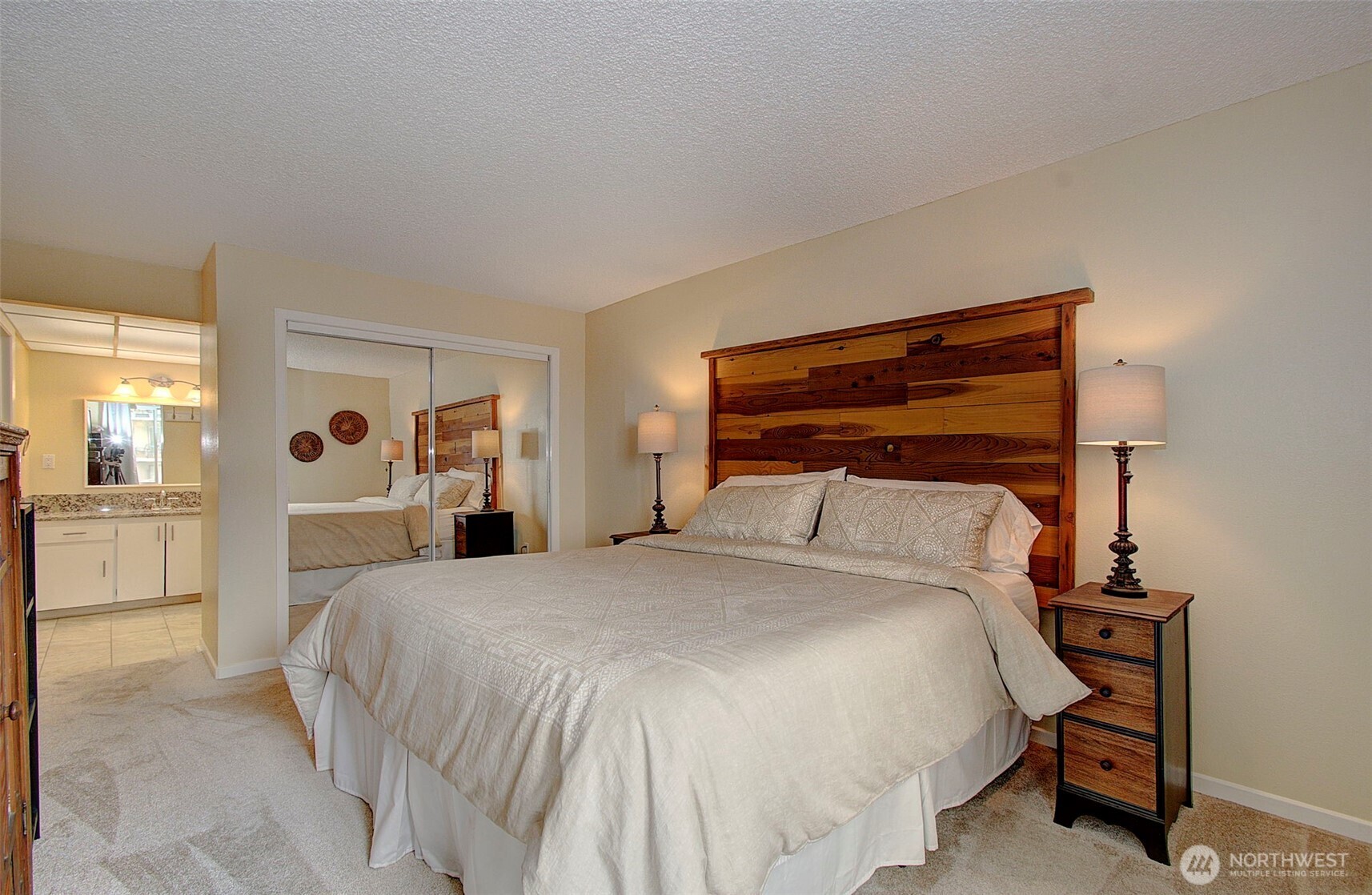Bedroom with a large bed, rustic wood headboard, mirrored closet doors, and an adjoining bathroom vanity.