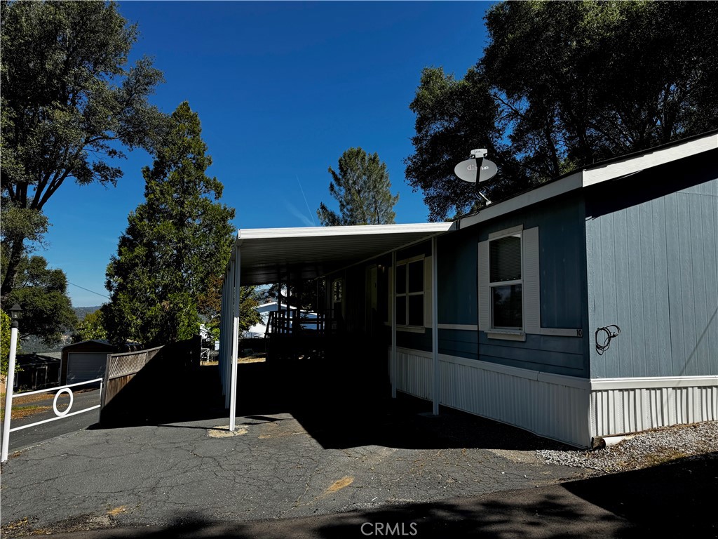 Blue manufactured home with white trim and a covered carport beneath mature trees.