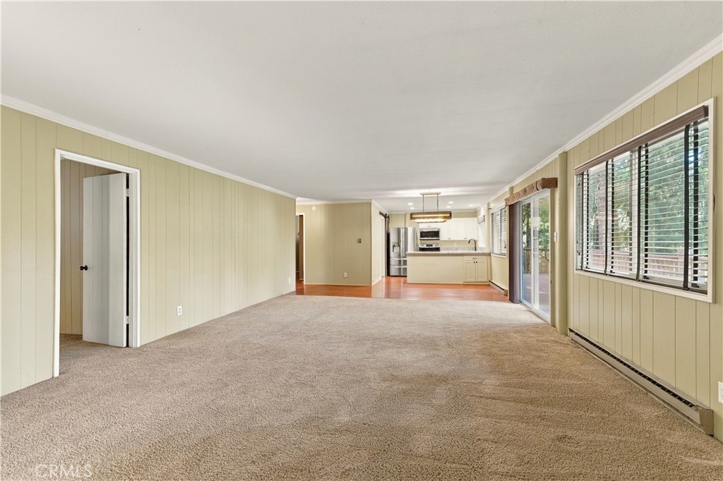 Unfurnished living room opening to a kitchen, with beige carpet and wide windows.