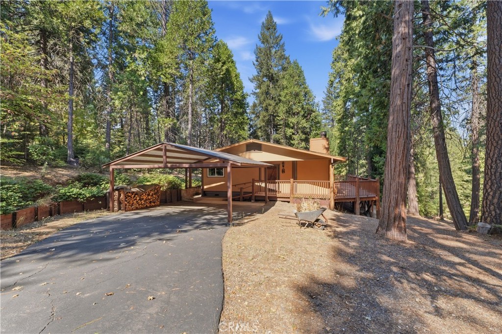 Wood-sided home among tall pines, with a covered carport, raised deck, and paved driveway.