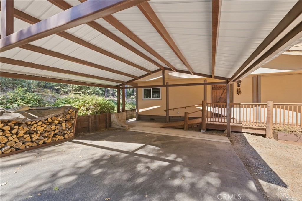 Covered carport with concrete parking pad, stacked firewood, and a wooden ramp to the house.